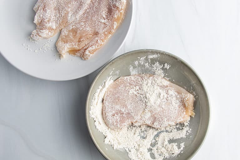 Overhead view of thin chicken cutlets in a shallow dish with a flour-based breading mixture.