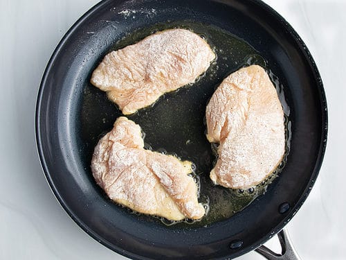 Overhead view of 3 lightly breaded chicken cutlets in a large non-stick skillet.