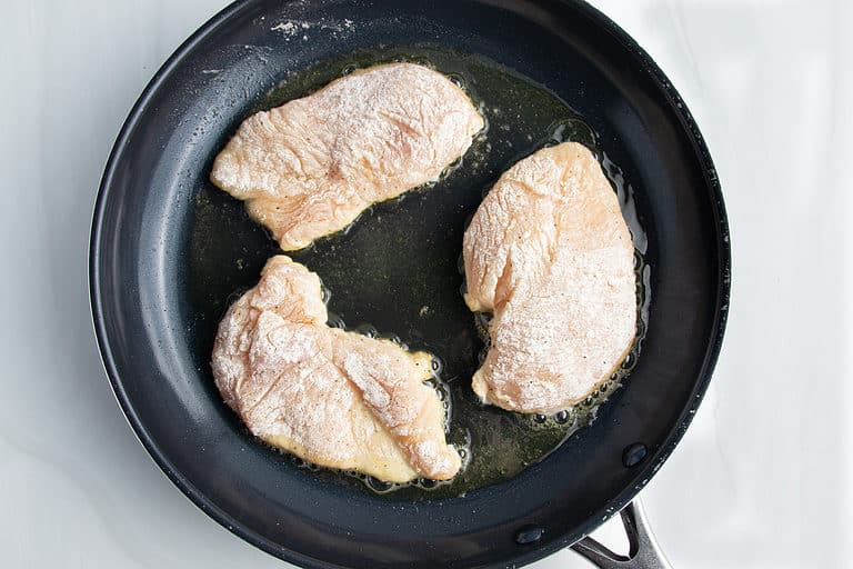 Overhead view of 3 lightly breaded chicken cutlets in a large non-stick skillet.