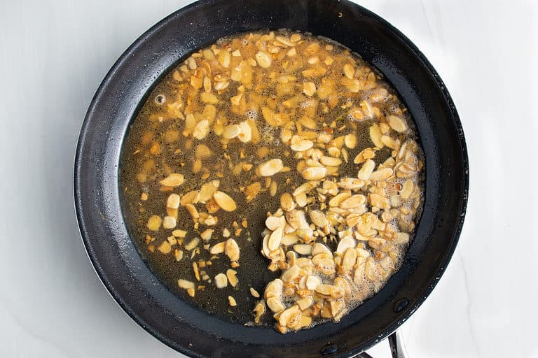 Overhead view of chicken almondine sauce (butter, chicken broth, lemon juice, and almonds) in a large black non-stick skillet.