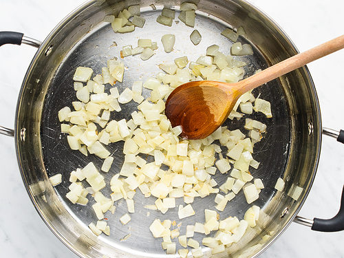 Overhead view of sautéed onion in a large silver skillet with a large wooden spoon on a white background.