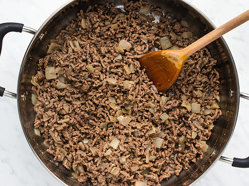 Overhead view of sautéed onions and crumbled ground beef in a large silver skillet on a white table with a wooden spoon.