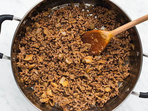 Overhead view of seasoned ground beef and sautéed onions in a large silver skillet with a wooden spoon on a white background.