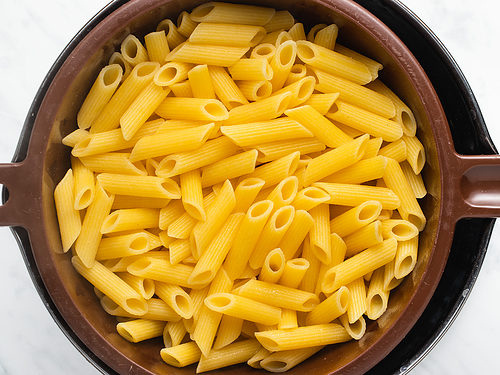 Overhead view of cooked penne pasta in a colander resting over a large pot on a white tabletop.