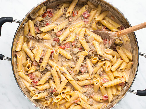 Overhead view of Rotel pasta with ground beef and a creamy sauce in a large silver skillet with 2 handles and a wooden spoon on a white tabletop.