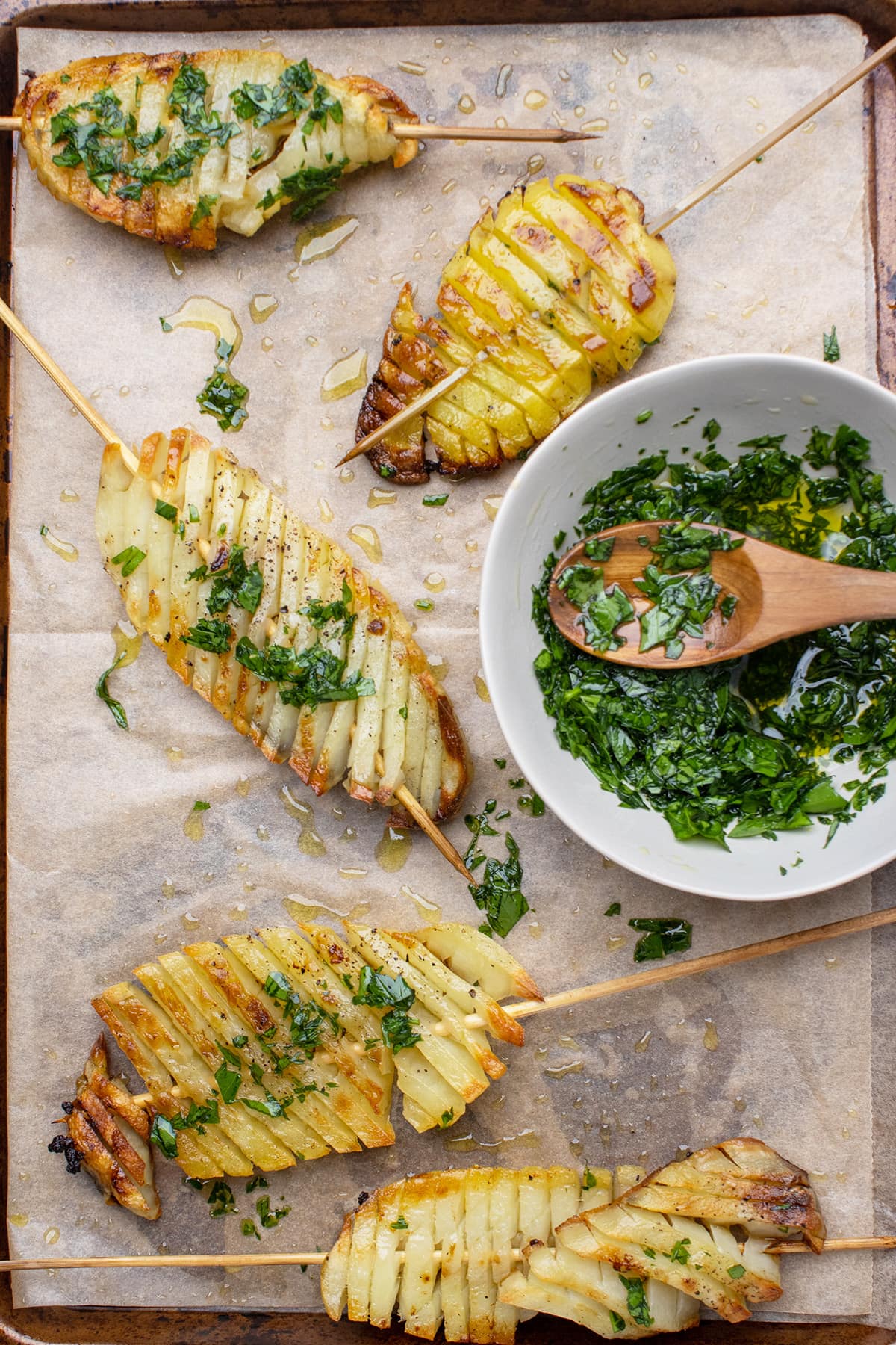 Overhead view of 5 accordion potato skewers on a baking sheet lined with parchment paper, next to a bowl of chimichurri sauce.