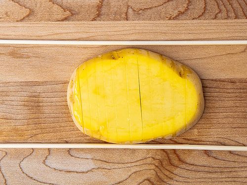 Slice of potato laying horizontally on cutting board with skewers on either side of potato.