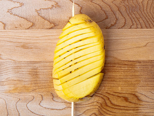 Overhead view of potato, sliced diagonally, skewered onto a wooden skewer, resting on a cutting board.