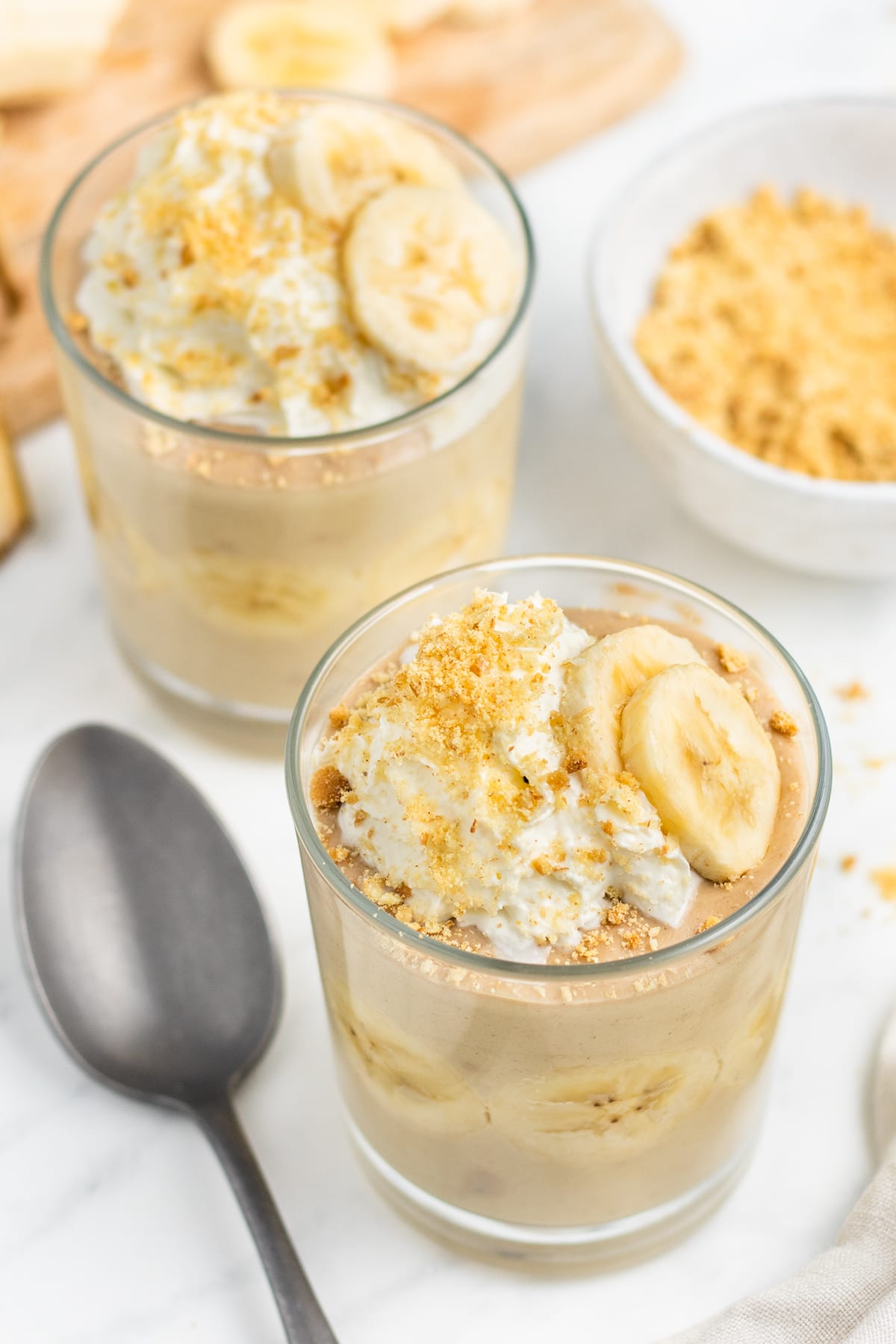 Overhead, angled view of two glass cups of 3-ingredient banana pudding topped with banana slices and whipped cream.
