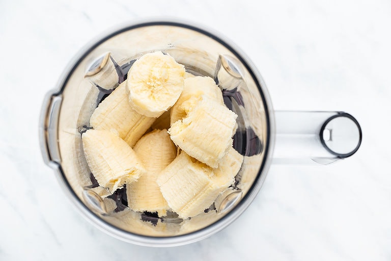 Overhead view of bananas in a blender on a white countertop.