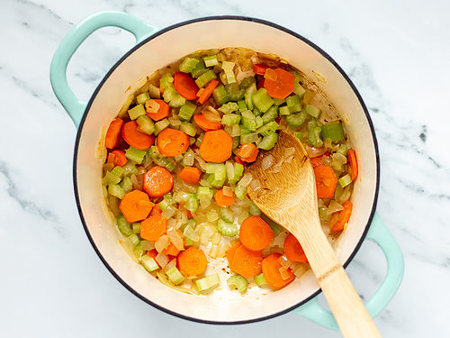 Olive oil, onions, garlic, chopped celery, and chopped carrots in a large soup pot with double handles and a wooden spoon.