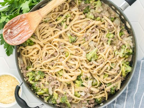 Ground beef alfredo with broccoli florets in a large skillet with a wooden spoon.
