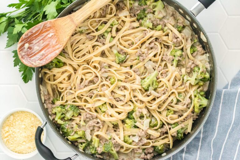 Ground beef alfredo with broccoli florets in a large skillet with a wooden spoon.