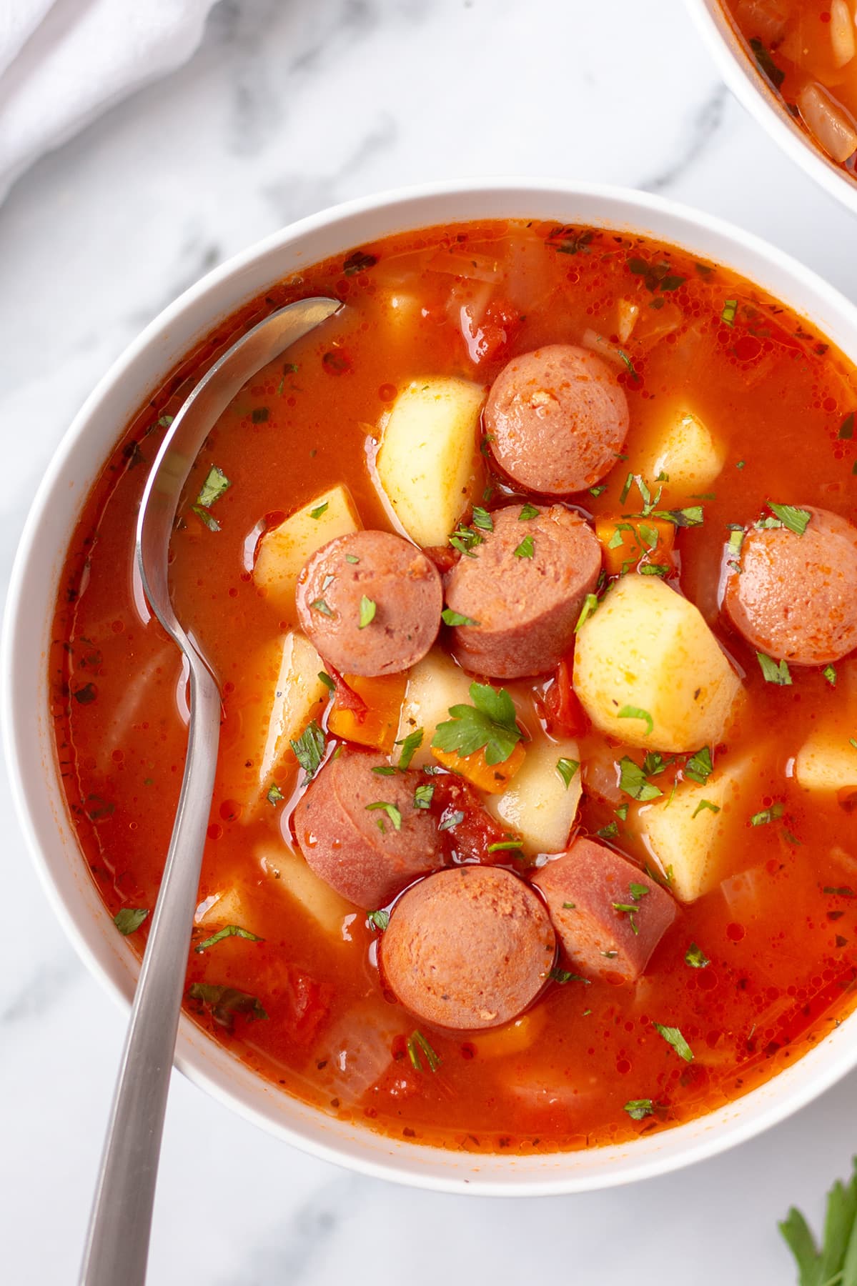 Close-up, overhead view of a white bowl of bright red hot dog soup on a white tablecloth.
