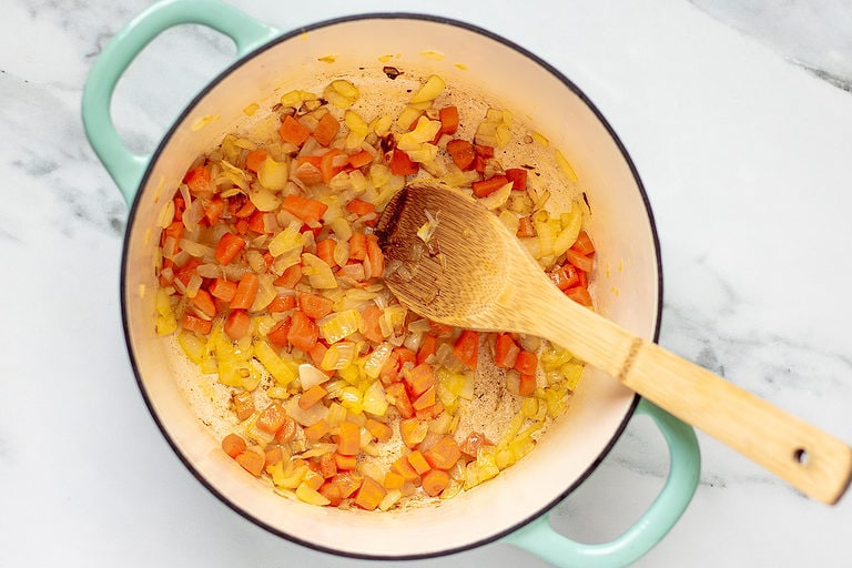 Overhead view of a large pot holding chopped onions, carrots, and garlic. A wooden spoon rests with the handle on the edge of the pot.