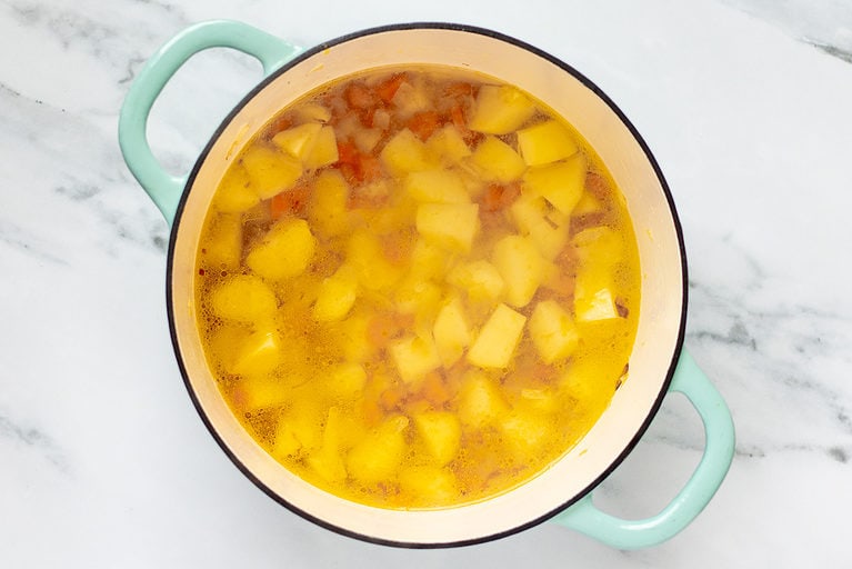 Overhead view of potatoes, water, carrots, onions, and garlic in a large soup pot on a marble countertop.
