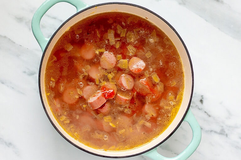 Overhead view of hot dog soup in a large soup pot on a white and grey marble countertop.