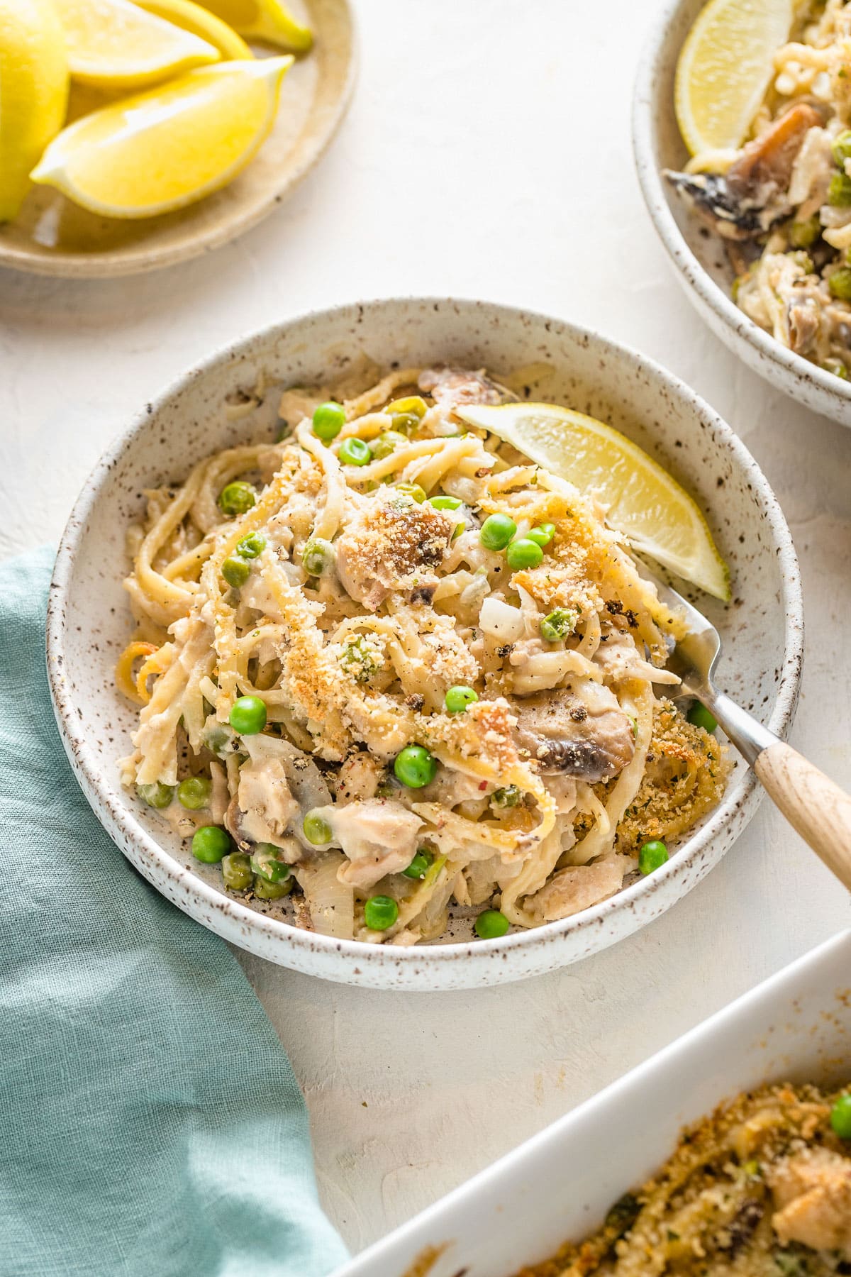 A white serving bowl with tuna tetrazzini, lemon wedges, and peas on a neutral table with a blue napkin.