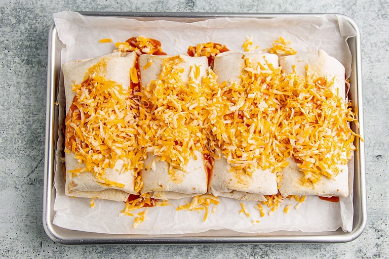 Rolled burritos topped with shredded cheese on a baking sheet.