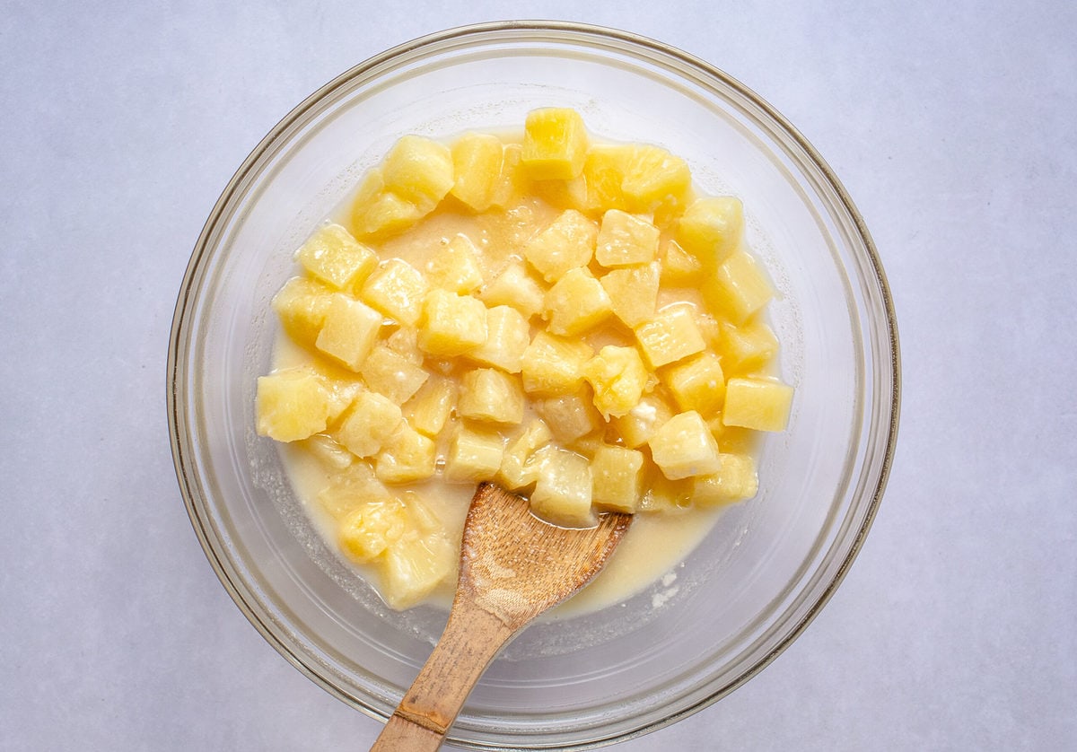 Pineapple casserole filling in a large glass mixing bowl with a wooden spoon.