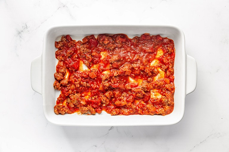 Red marinara meat sauce covering bottom of white rectangular baking dish.