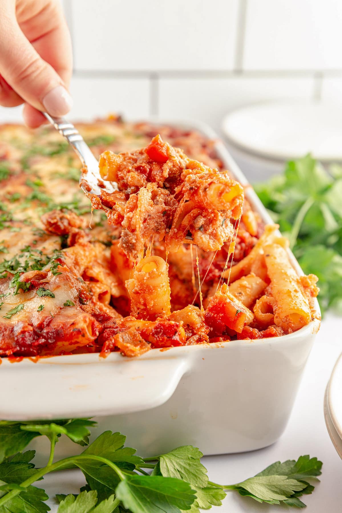 Baked ziti pasta being scooped out of a casserole dish by a white woman's hand holding a silver spoon.