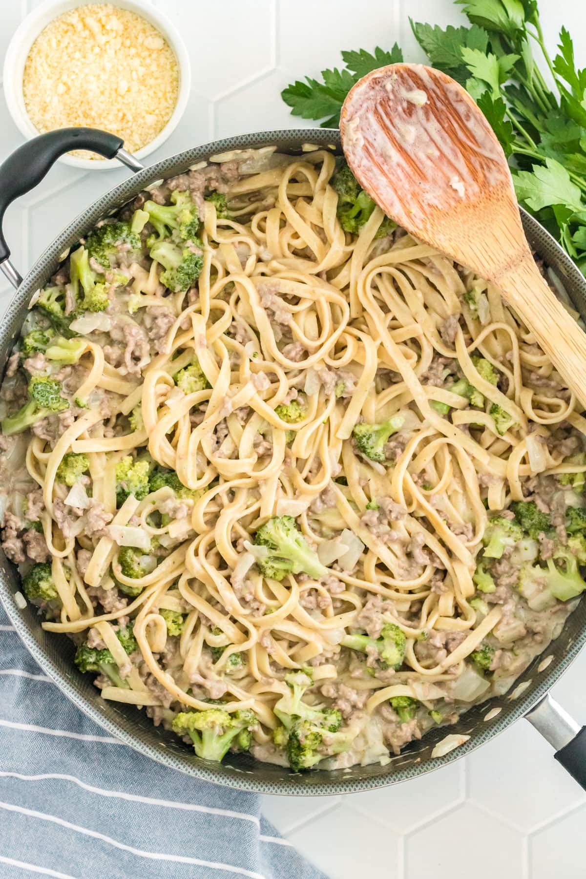 Ground beef alfredo with broccoli florets in a large skillet with a wooden spoon.