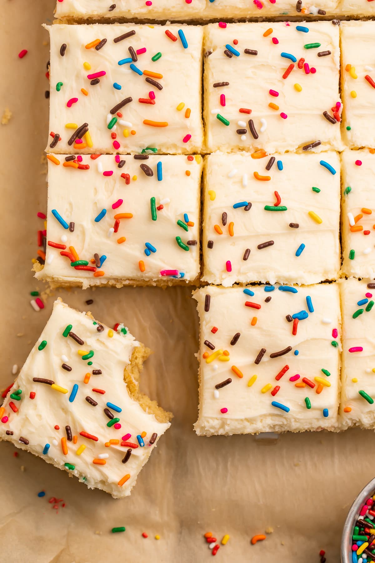 Overhead view of a dozen vanilla brownies topped with butter cream and sprinkles. One bite is missing from the bottom left brownie.