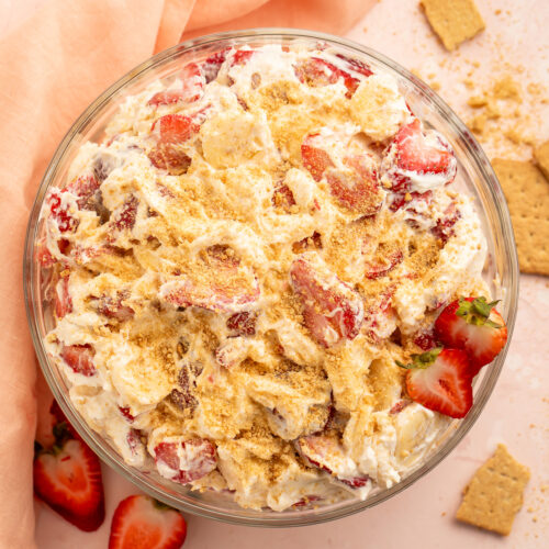 Top down view of a large bowl of creamy, thick strawberry cheesecake salad topped with crushed graham crackers on a pink tabletop with a pink cloth napkin.