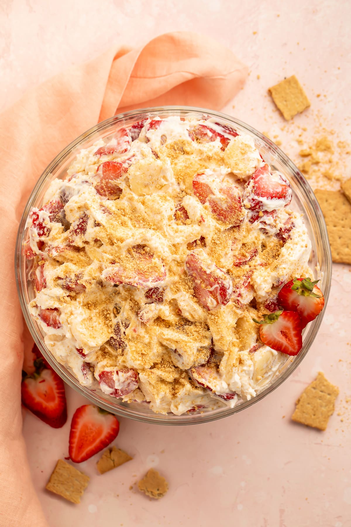 Top down view of a large bowl of creamy, thick strawberry cheesecake salad topped with crushed graham crackers on a pink tabletop with a pink cloth napkin.