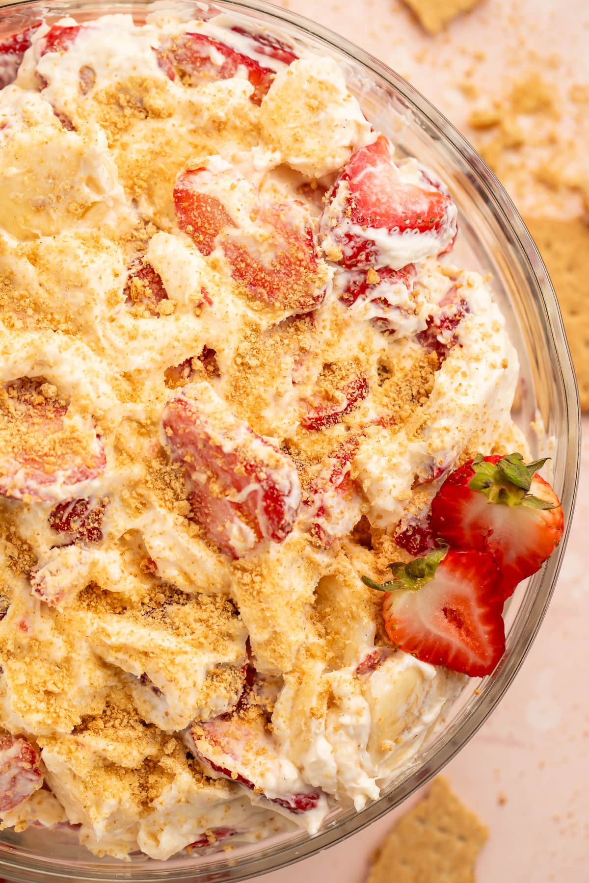 A large glass mixing bowl filled with thick, creamy cheesecake salad topped with sliced strawberries and bananas and crumbled graham crackers.