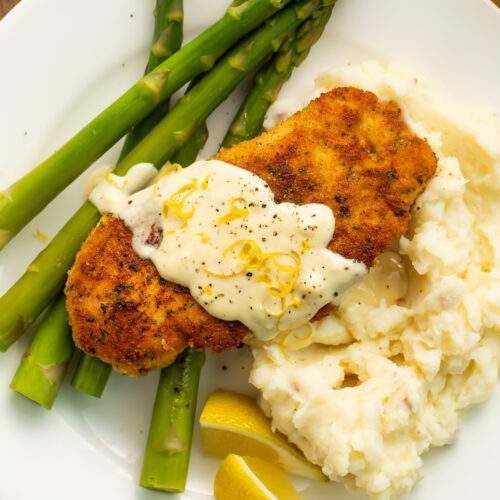 Breaded chicken costoletta topped with a cream sauce on a white plate with bright green asparagus.