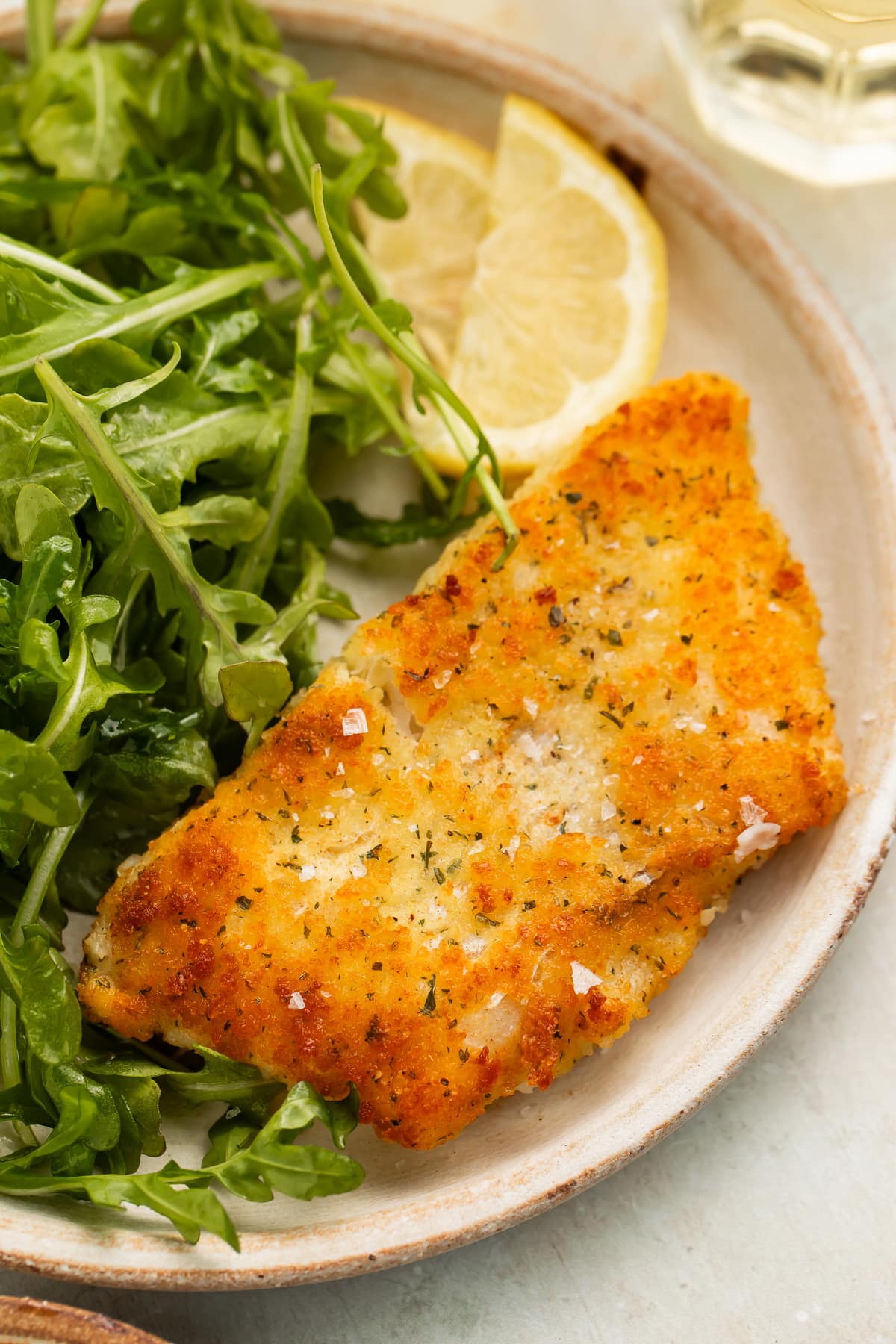 A fillet of potato crusted cod on a neutral plate with a small green salad and a wedge of lemon.
