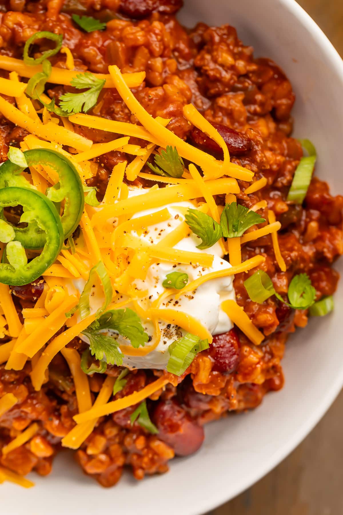 Close-up of a bowl of chili with rice, topped with shredded cheddar, sliced jalapeños, and a dollop of sour cream.