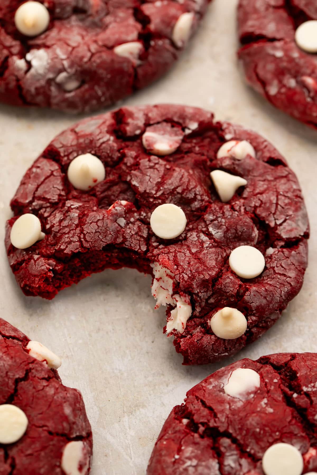 A crinkly, soft red velvet cake mix cookie dotted with white chocolate chips on a sheet of parchment paper. One bite has been taken out of the cookie.