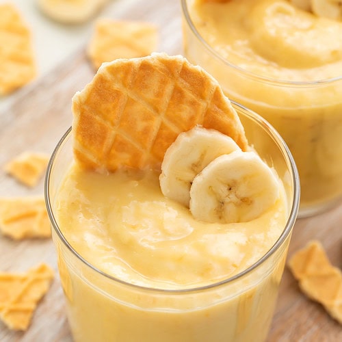 A small glass cup holding banana custard with a waffle cone cookie and two slices of banana on top for garnish.