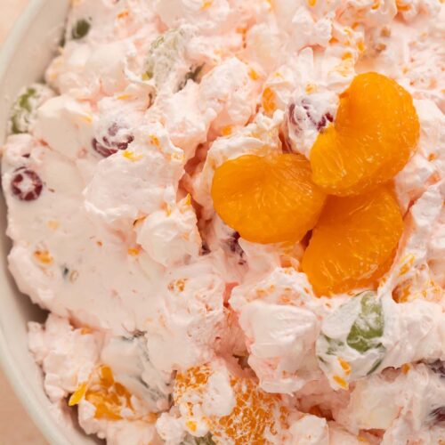 A large bowl of fruit salad made with Cool Whip, maraschino cherries, mandarin oranges, seedless grapes, and marshmallows.