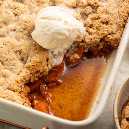 A baking dish of sweet potato cobbler, with a large scoop removed from the corner of the dish, revealing the sweet potato slices beneath the cobbler topping.