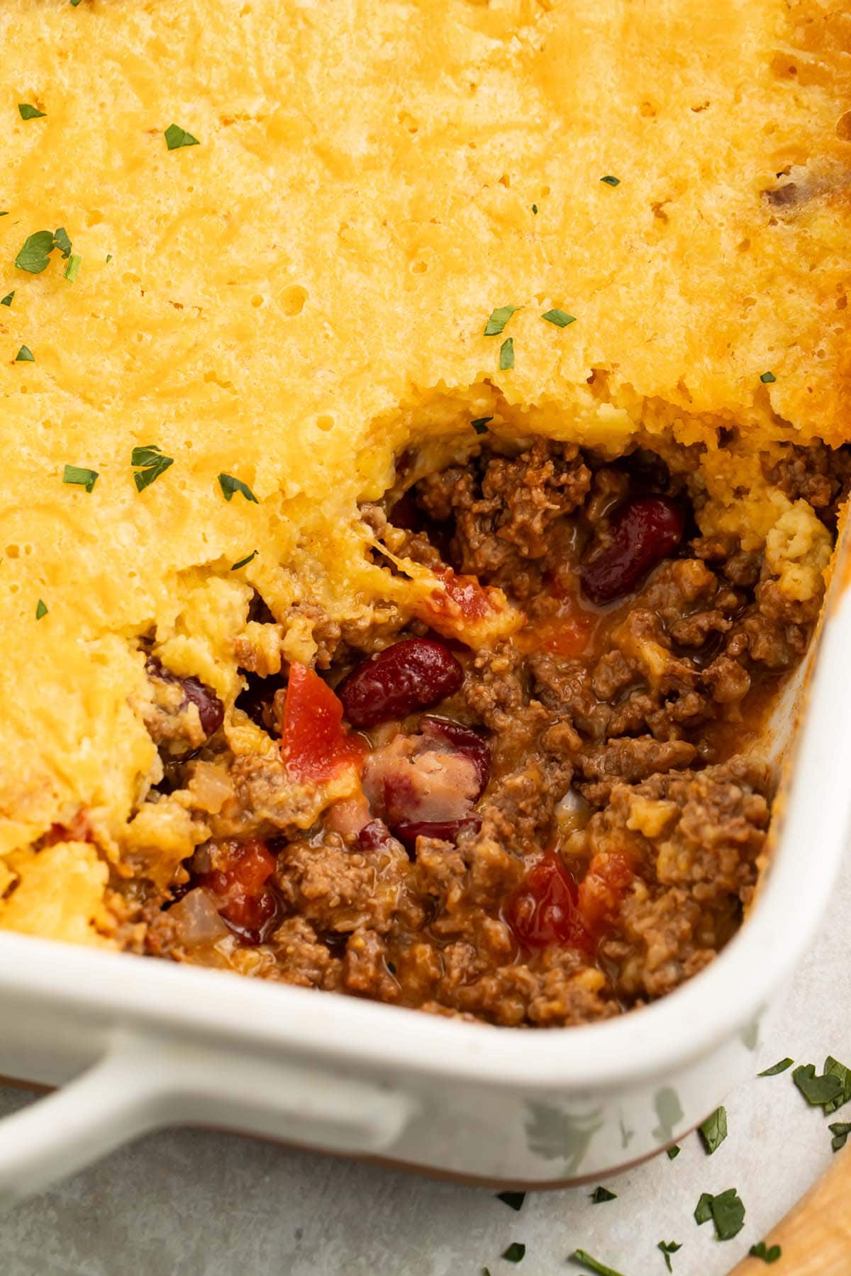 A large white casserole dish holding cowboy cornbread casserole, with a large scoop missing from the front corner, showing the ground beef and bean mixture under the cornbread topping.