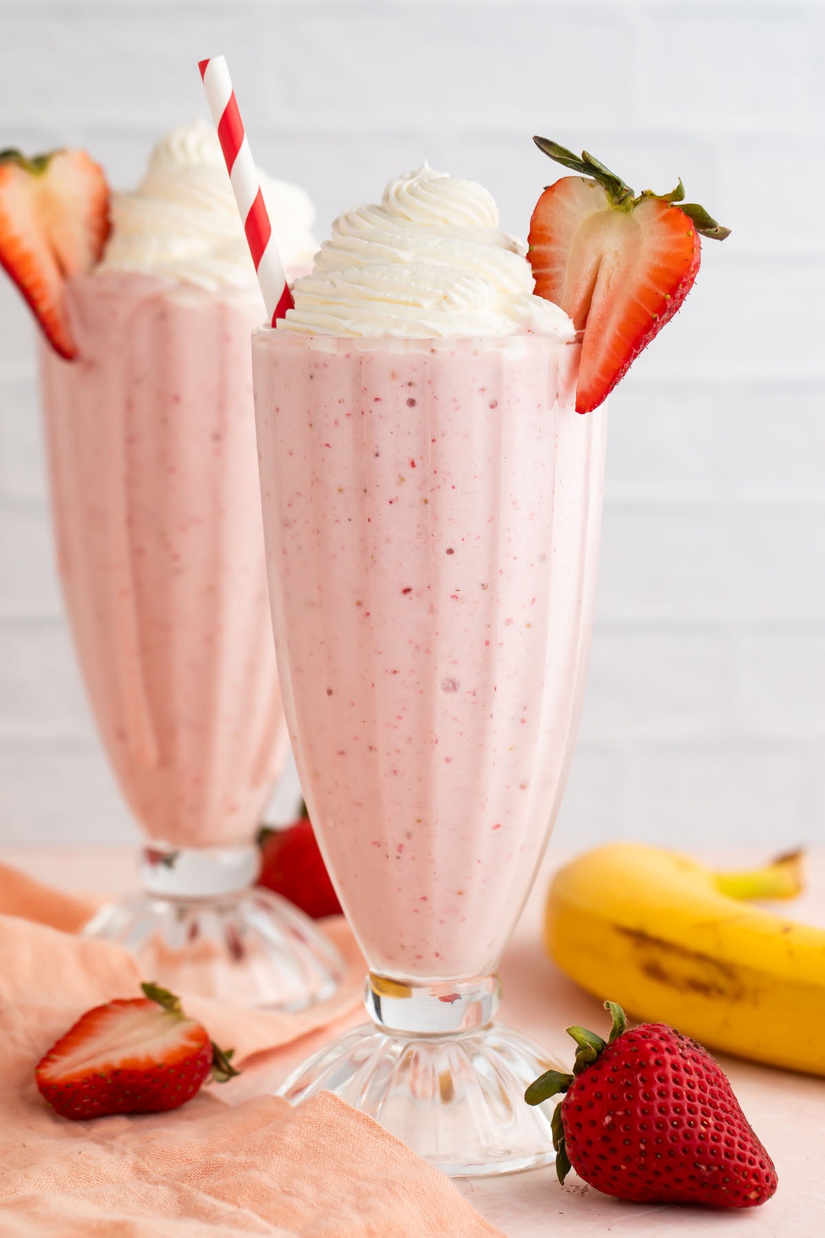 Two retro-style glasses containing light pink strawberry banana milkshakes topped with whipped cream and garnished with a half strawberry on the rim.