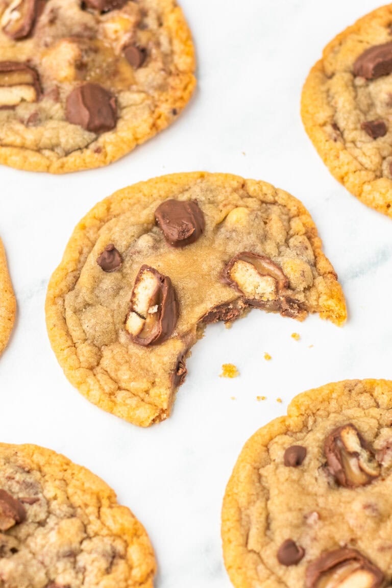 A Twix cookie with one bite missing, laying on a sheet of parchment paper surrounded by other cookies.