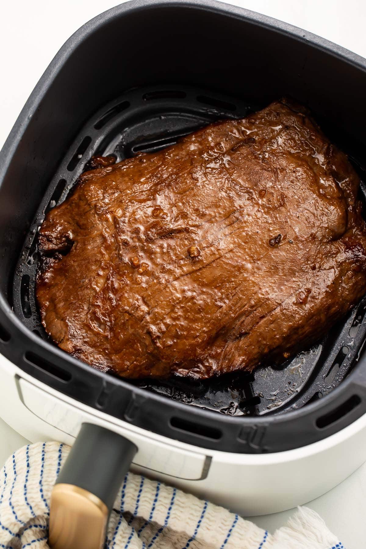 A whole, cooked flank steak resting in an air fryer basket on a counter top.