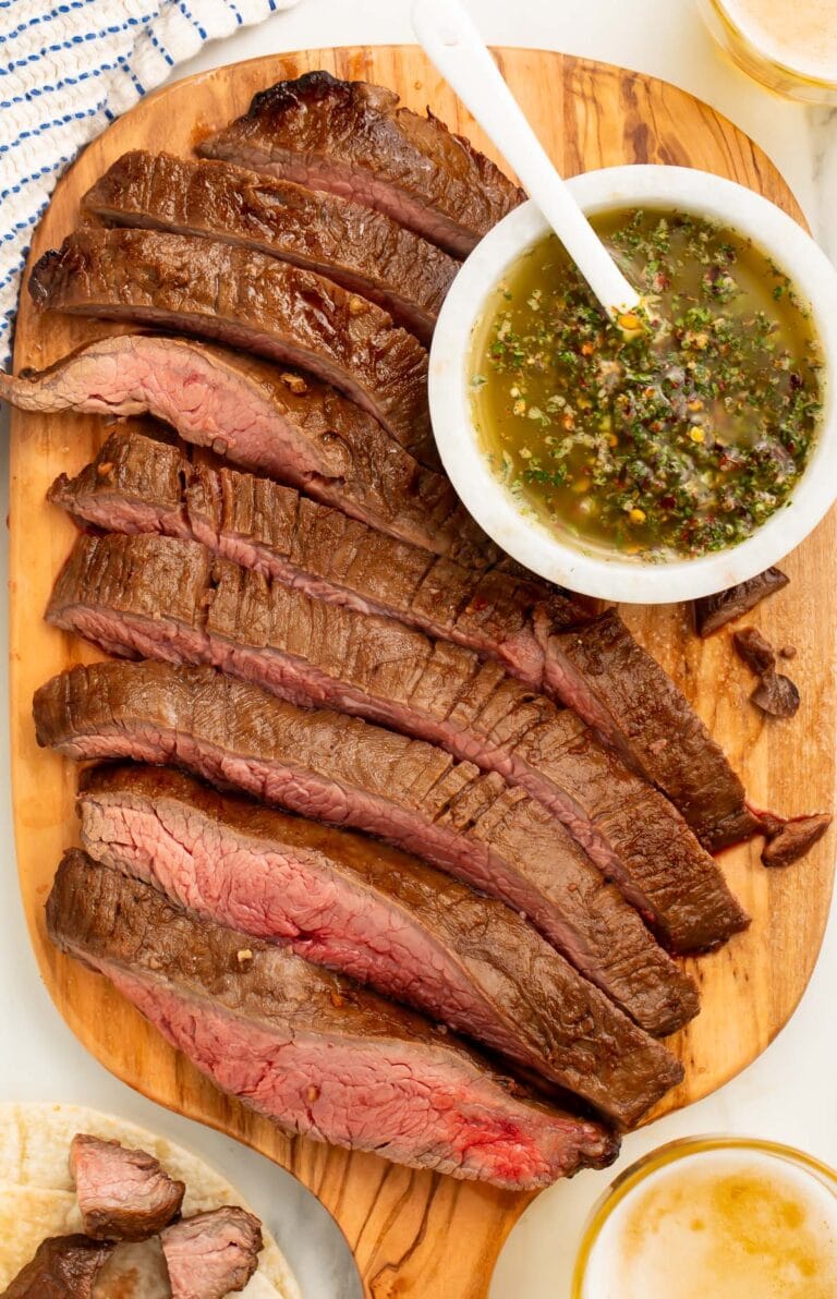 Air fryer flank steak, sliced into strips and arranged on a wooden serving board next to a bowl of chimichurri sauce.
