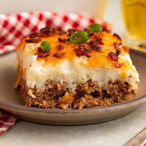Slice of cowboy meatloaf and potato casserole on a plate with the rest of the casserole and a glass of beer in the background.