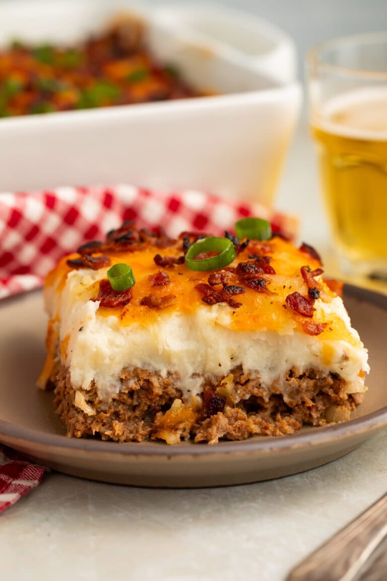 Slice of cowboy meatloaf and potato casserole on a plate with the rest of the casserole and a glass of beer in the background.