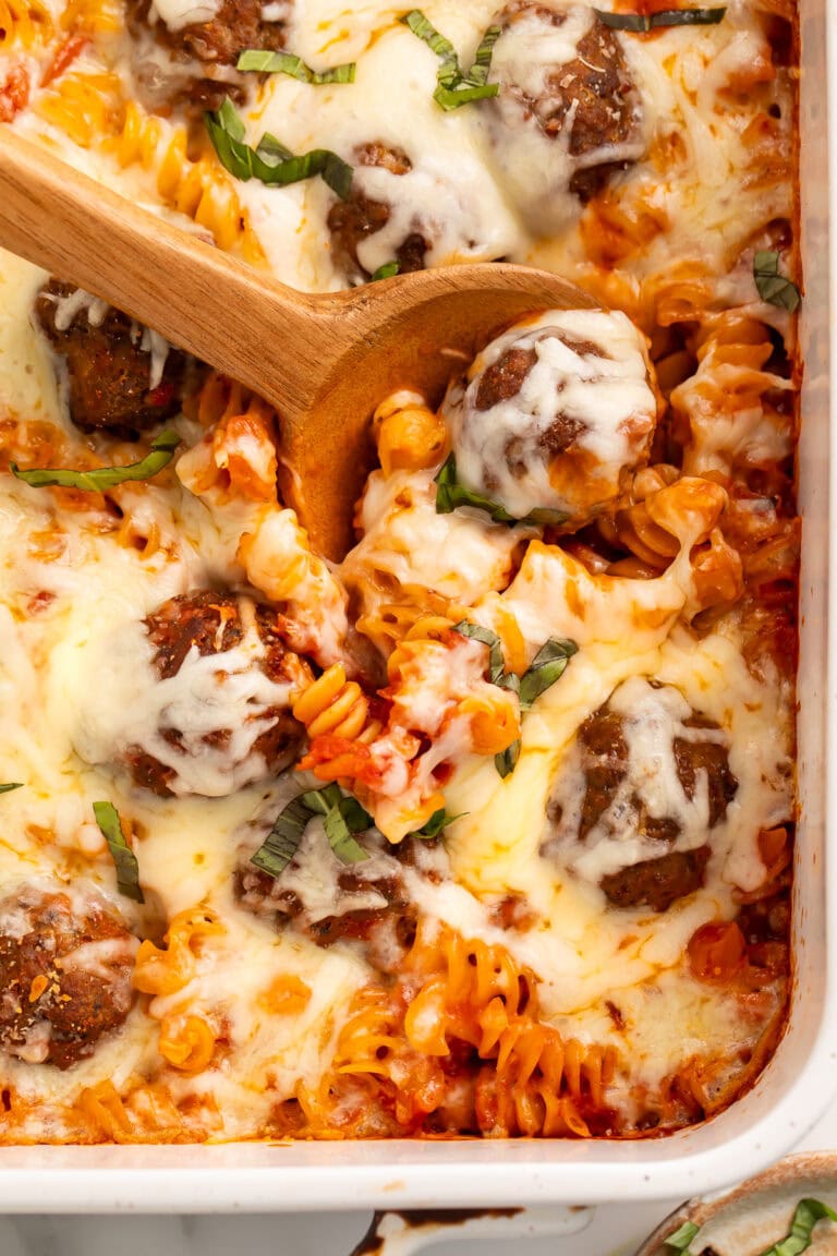 Wooden spoon serving a meatball pasta bake in a white baking dish.