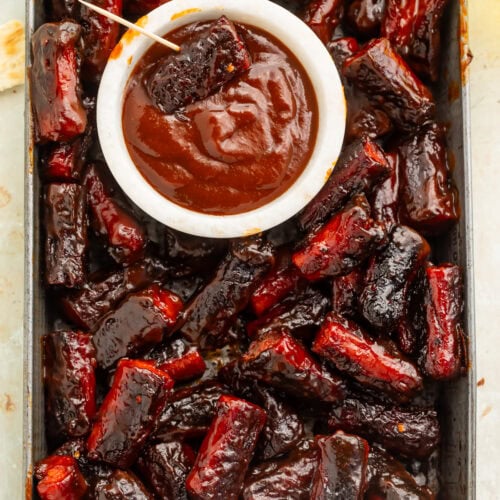 Metal fray of hot dog burnt ends with a white pot of barbecue sauce near the middle for dipping.