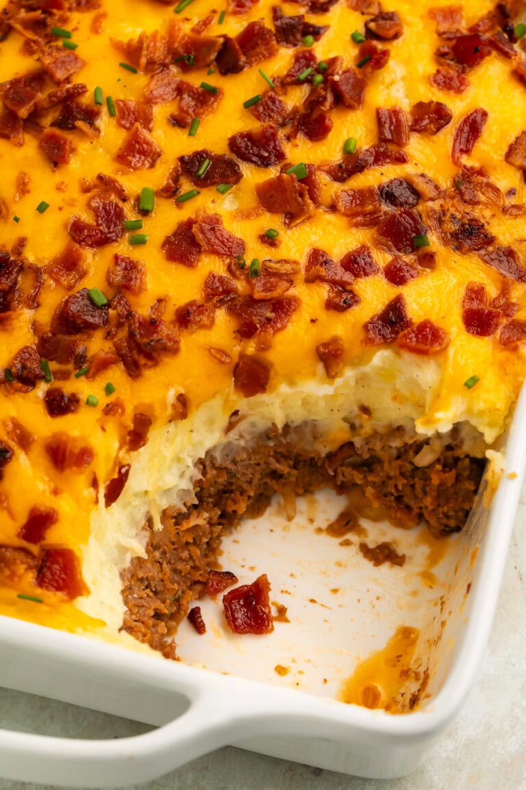 Bacon, cheese and chive-topped mashed potato meatloaf casserole with a spoonful taken out of the corner to show the meat layer underneath.