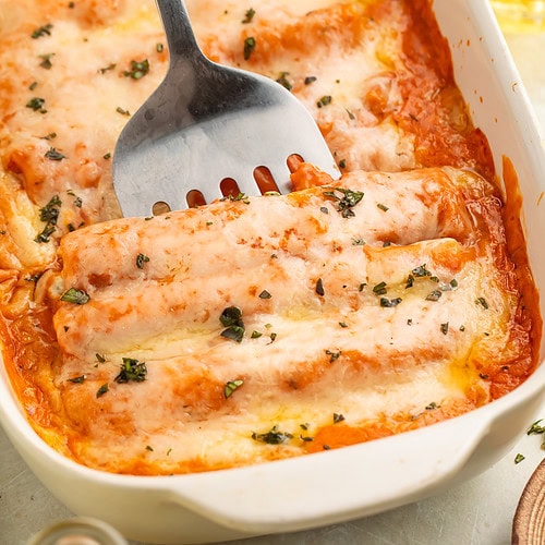 A metal kitchen tool being slide into a pan of cooked chicken canelloni topped with a creamy tomato sauce and melted cheese.
