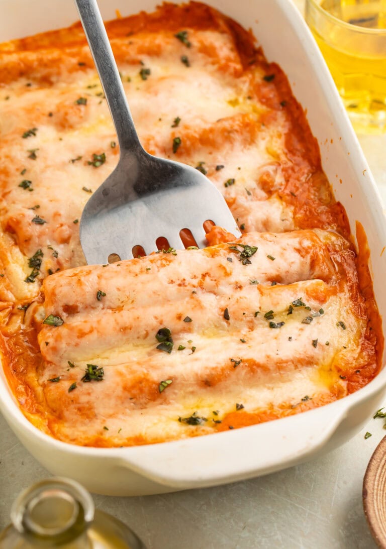 A metal kitchen tool being slide into a pan of cooked chicken canelloni topped with a creamy tomato sauce and melted cheese.